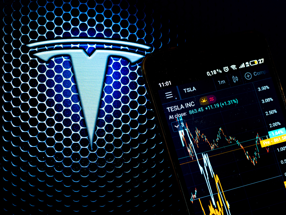 investicni aplikace na mobilu sledujici vyvoj cen akcie tesly a v pozadi logo automobilky tesla